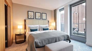 Quarto de hotel com uma cama grande, duas mesas de cabeceira, um banco e vista para um edifício de tijolos através de uma janela ampla.