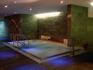 Una piscina interior moderna con un fondo de pared de piedra, una escalera metálica y un banco integrado.