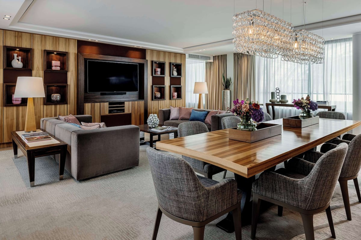 A modern living room with a large flat-screen TV, comfortable seating, a dining area with a wooden table and chairs, and a decorative chandelier.