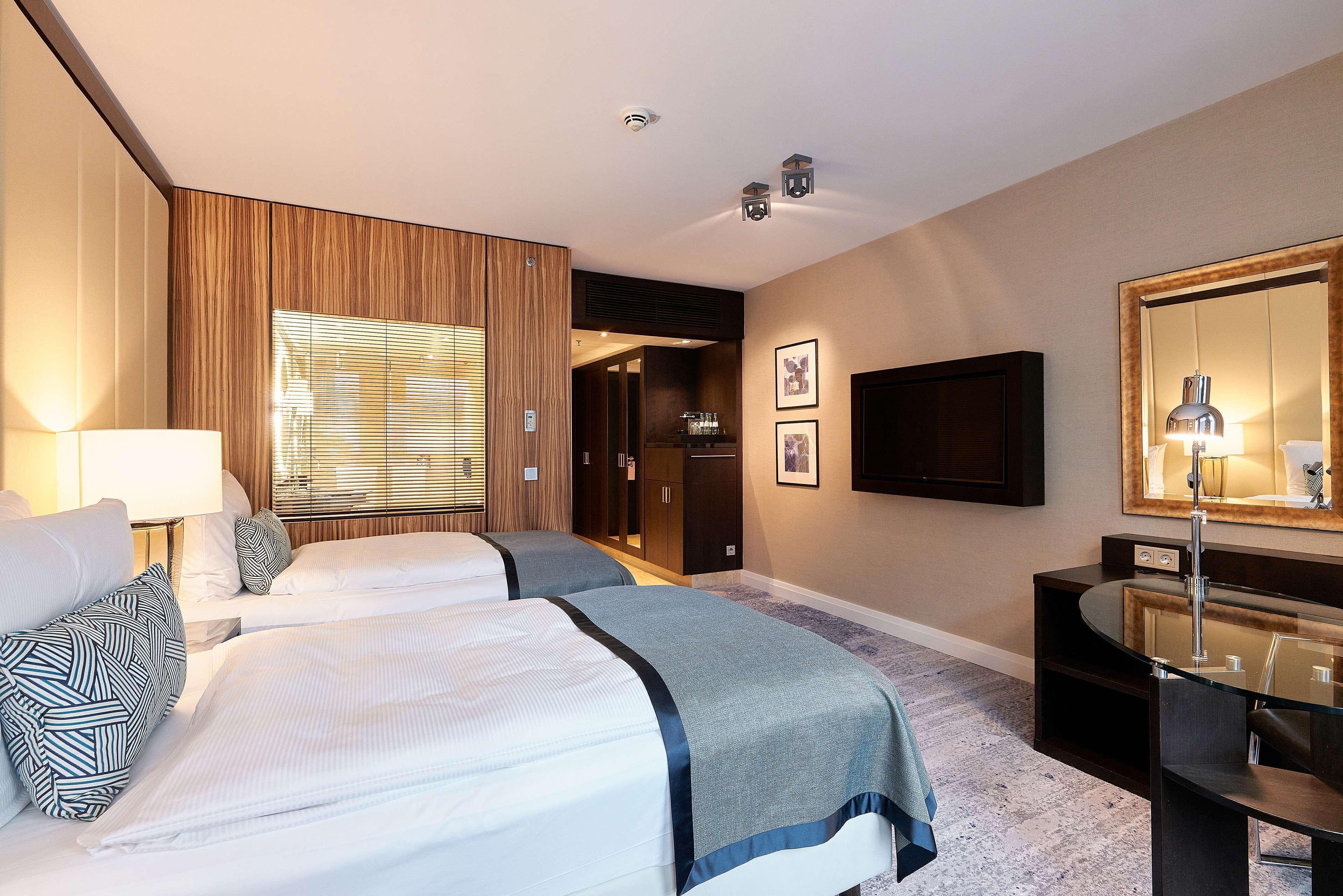 A hotel room with two beds, a TV, a desk with a mirror, and a window with blinds.