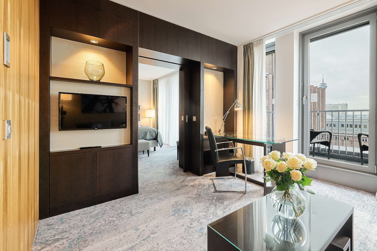 A modern hotel room with a large window, a desk with a vase of flowers, a flat-screen TV, and a seating area with a sofa and chairs.