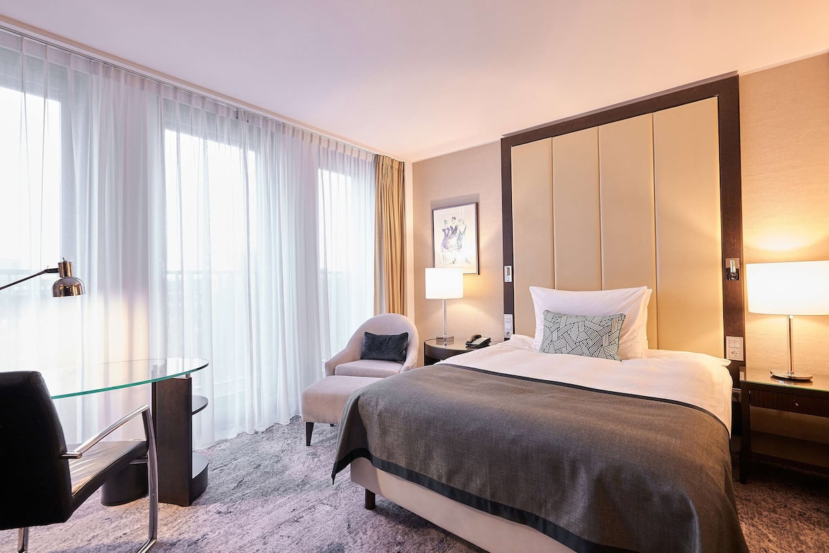 A hotel room with a large bed, a desk with a chair, a bedside table with a lamp, and a large window with curtains.