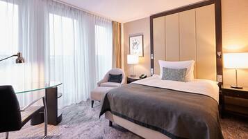 A hotel room with a large bed, a desk with a chair, a bedside table with a lamp, and a large window with curtains.