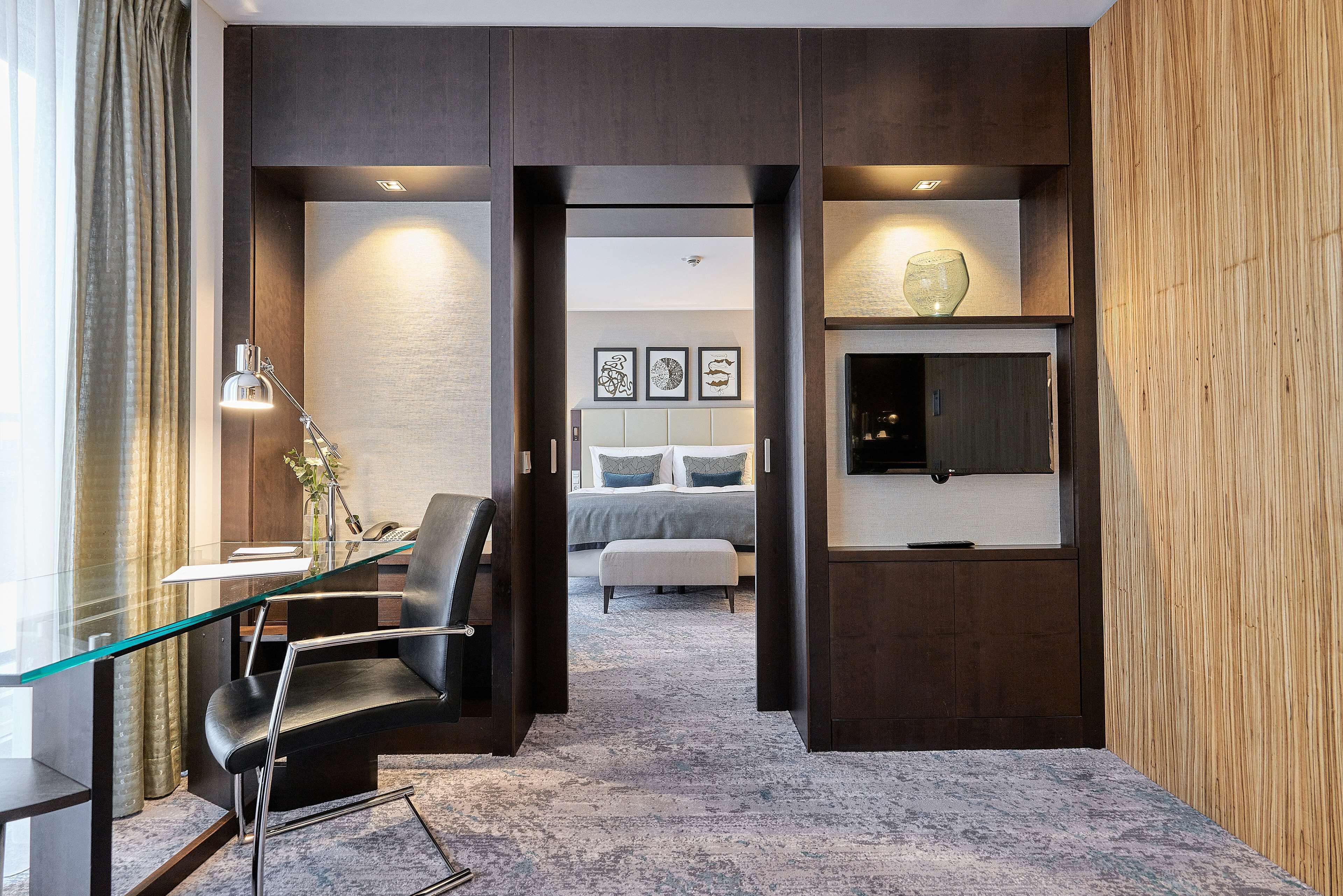 Ein modernes Hotelzimmer mit Glastisch, schwarzem Ledersessel, Bett mit grau-weißer Bettdecke, montiertem Fernseher und gerahmten Kunstwerken an der Wand.