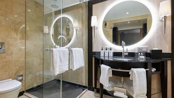 Een moderne badkamer met een douche met glazen wand, een ronde spiegel met ingebouwde verlichting, een zwart aanrechtbakje en een glazen plank met handdoeken.