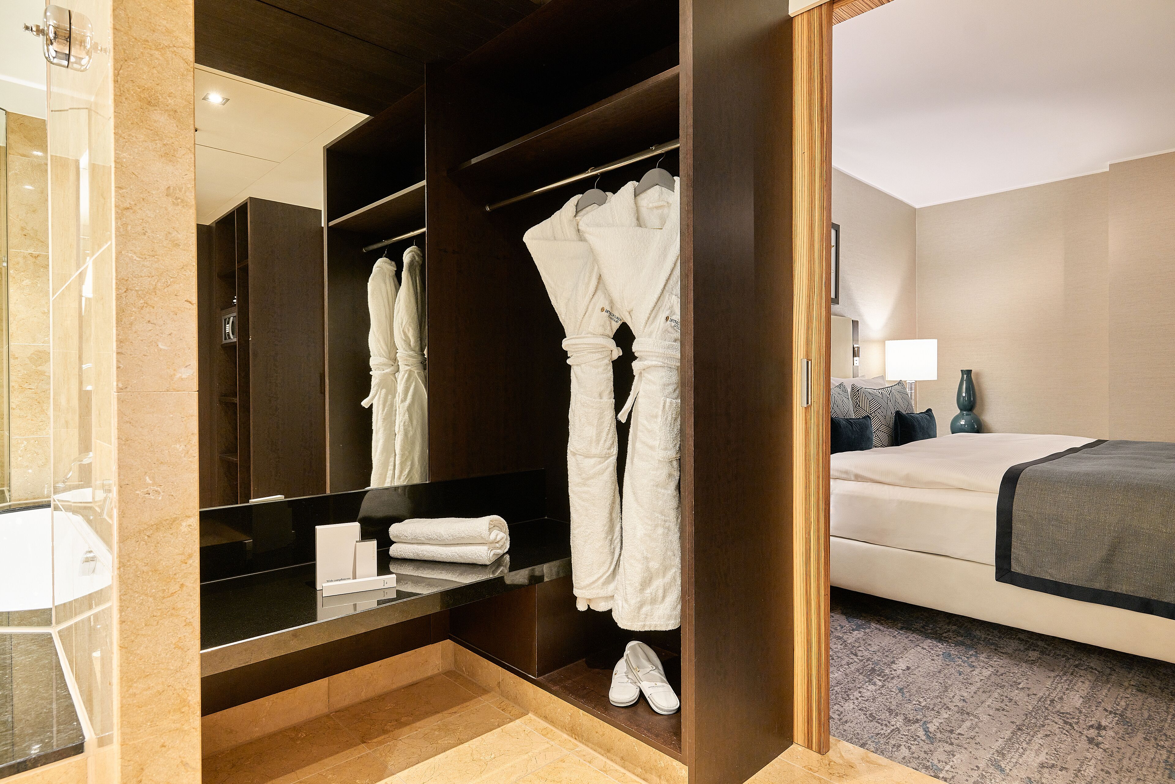 Habitación de hotel con vestidor, cama, mesita de noche y baño con lavabo y espejo.