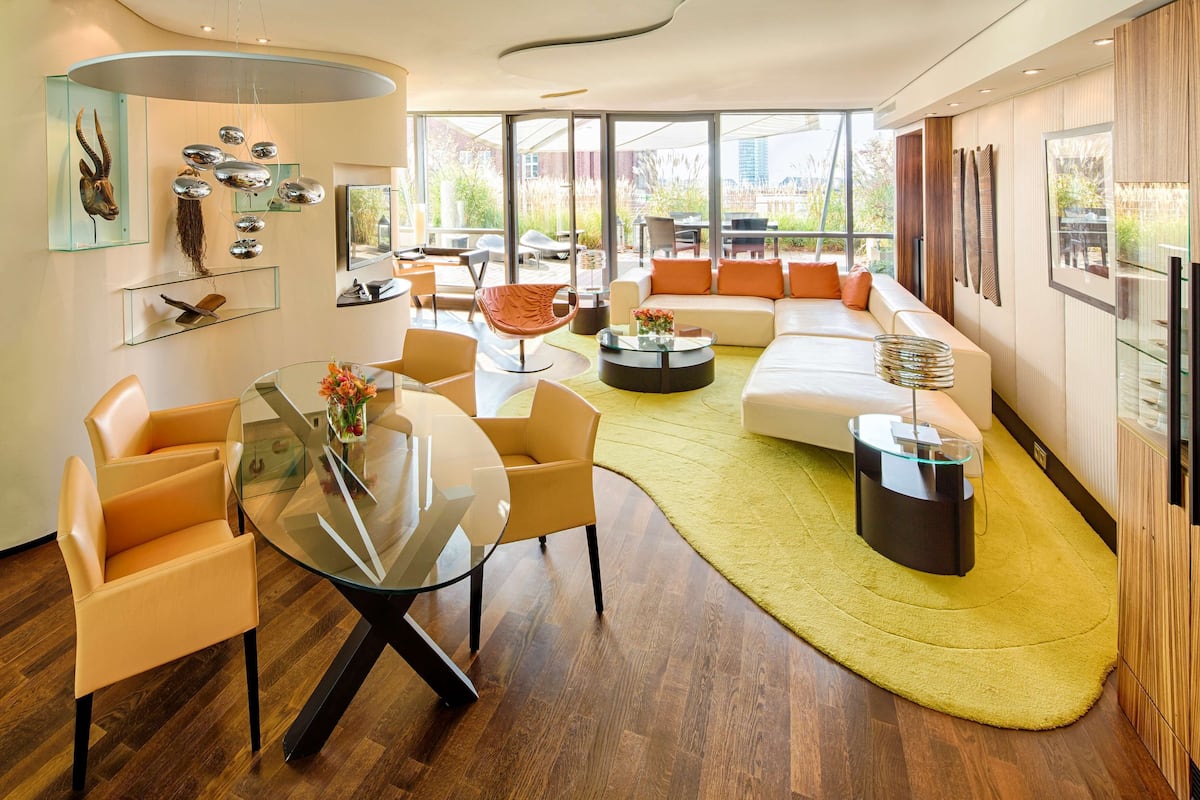 A modern living room with a glass dining table, comfortable seating, and a large window offering a view of the outdoors.