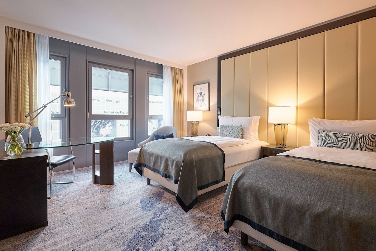 A hotel room with two beds, a desk, a chair, and a large window.