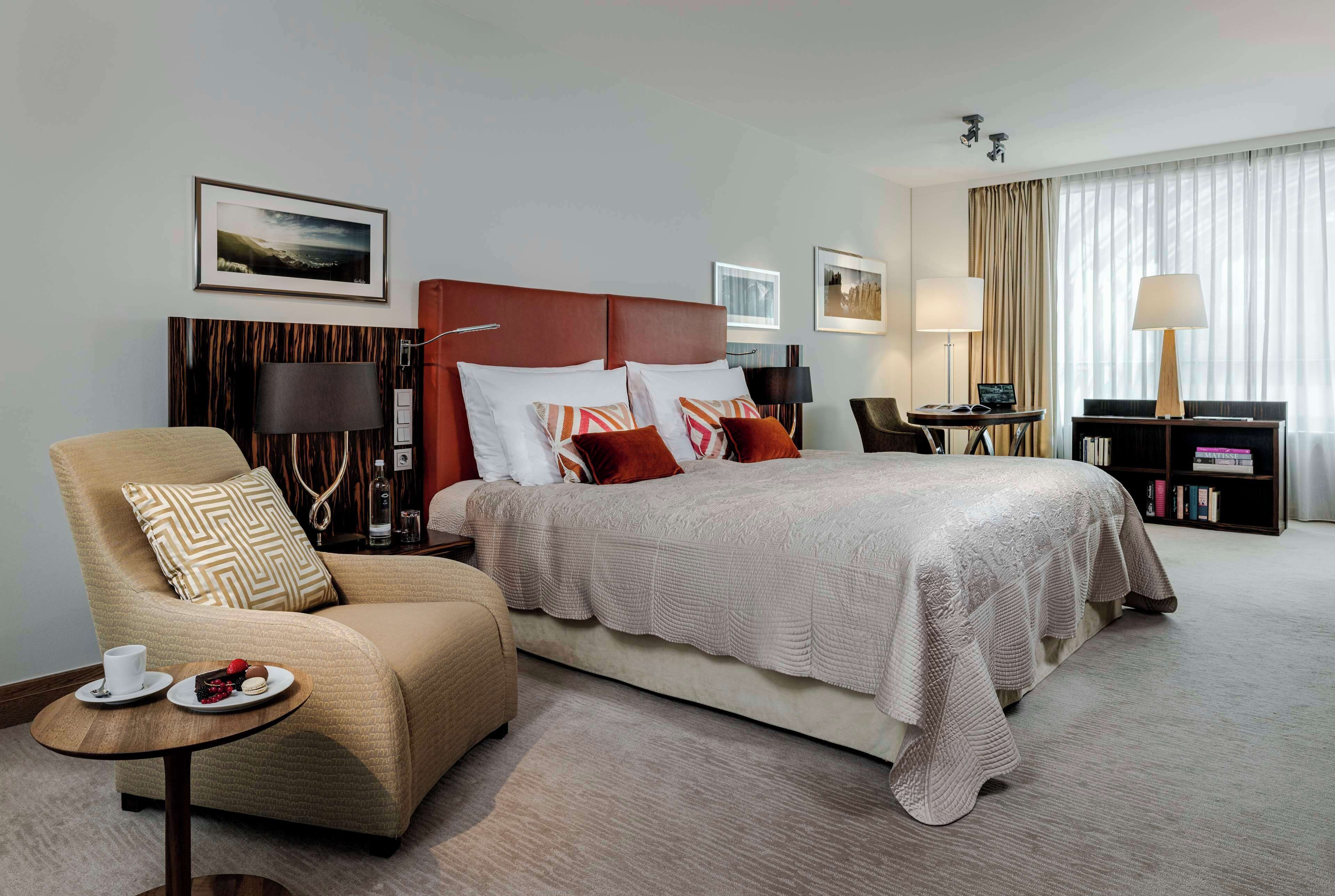 Une chambre d’hôtel avec un grand lit, un canapé, une petite table avec une tasse et des pâtisseries, une bibliothèque et un bureau avec une lampe.
