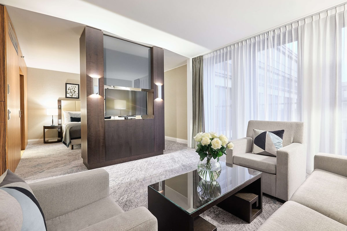 A modern hotel room with a sofa, armchair, and a glass coffee table. There is a large mirror and a flat-screen TV mounted on the wall.