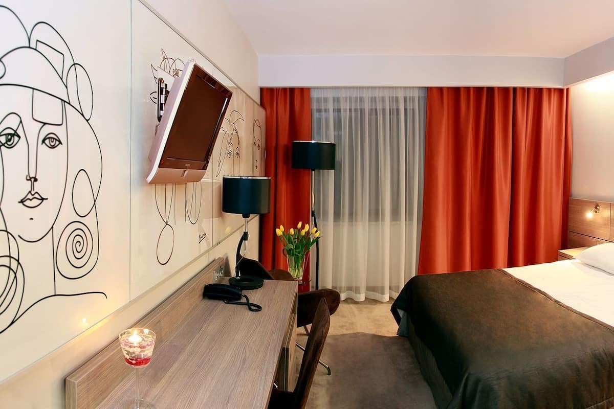 Ein Hotelzimmer mit Bett, Nachttisch, Lampe und einer Wand mit abstrakter Kunst.