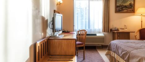 Una habitación de hotel con cama, escritorio con computadora, silla, ventana con cortinas y un cuadro en la pared.