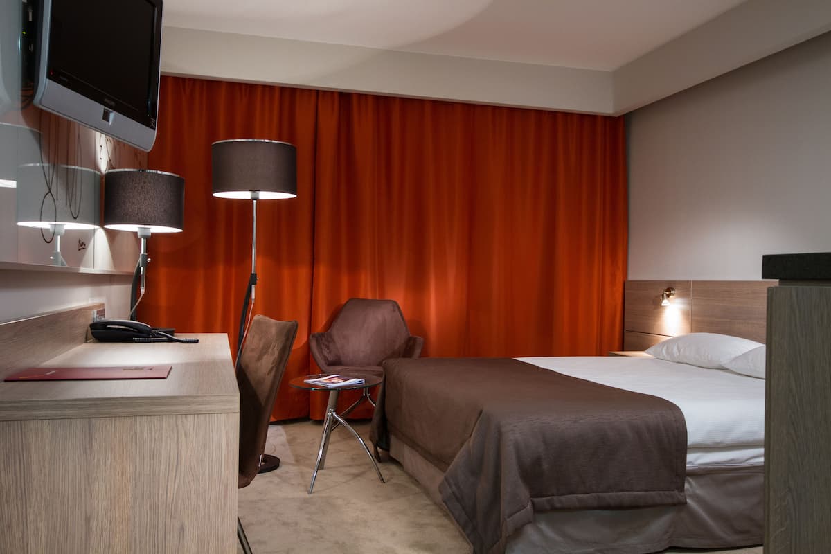 Ein Hotelzimmer mit einem Bett, einem Schreibtisch mit Telefon, einem Sessel, einem kleinen Tisch mit Lampe und einem an der Wand befestigten Fernseher.