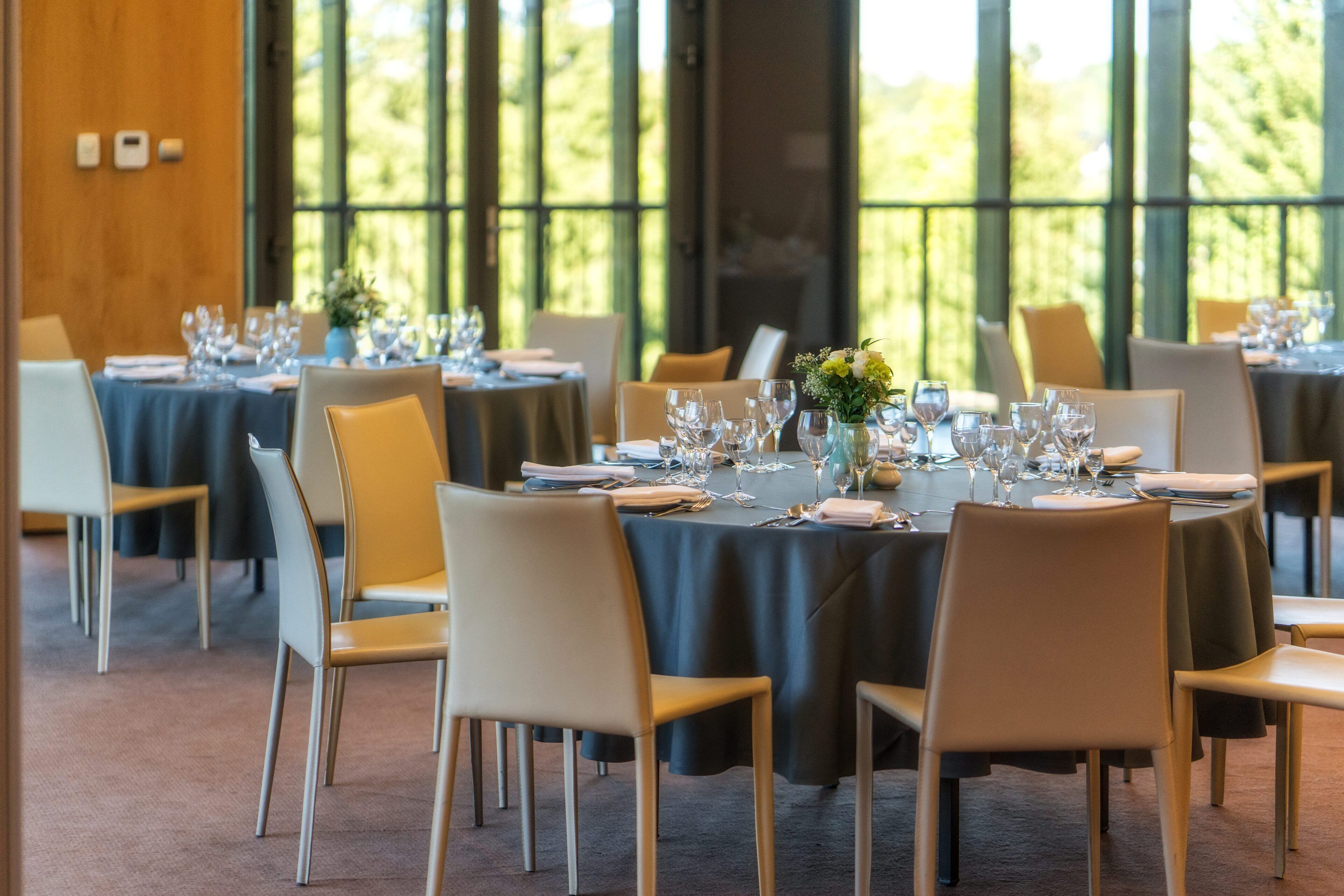 A dining area with round tables set for a meal, chairs, and a view of greenery through large windows.