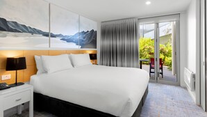 A modern hotel room with a large bed, a nightstand, a desk, and a view of a balcony with greenery. - Scenic Suites Queenstown (Queenstown)