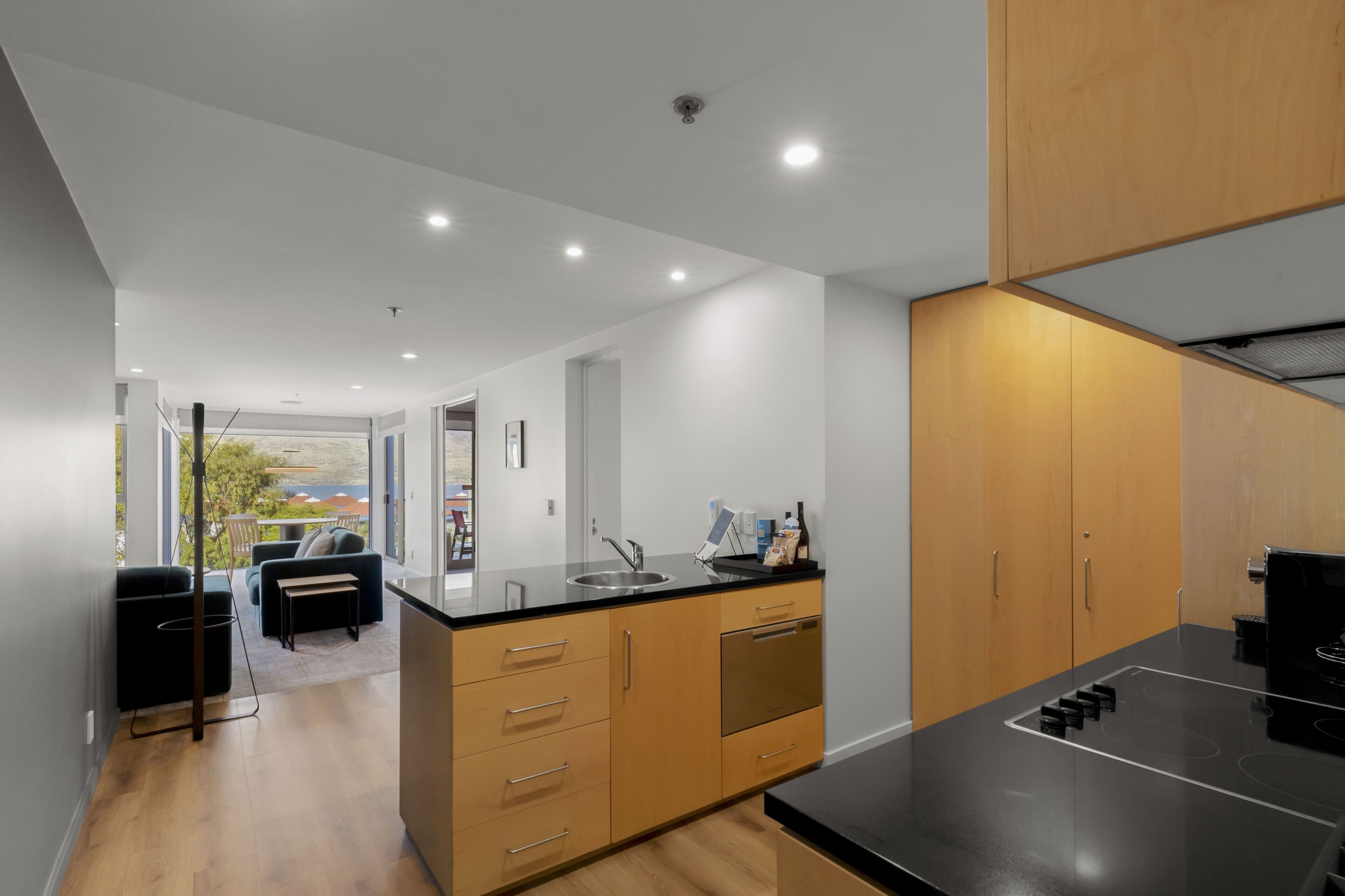 Una cocina moderna con encimera negra, armarios de madera y horno empotrado. Cuenta con una zona de comedor con mesa y sillas, y vistas al exterior a través de amplios ventanales.