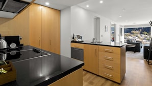 A modern kitchen with a black countertop, wooden cabinets, and a dishwasher. There is a view of a cityscape through a large window. - Scenic Suites Queenstown (Queenstown)