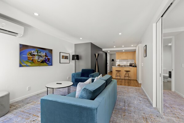 Una sala moderna con un sofá azul, una mesita redonda pequeña y un televisor montado en la pared. Se ve una cocina al fondo con gabinetes de madera y un taburete de bar.