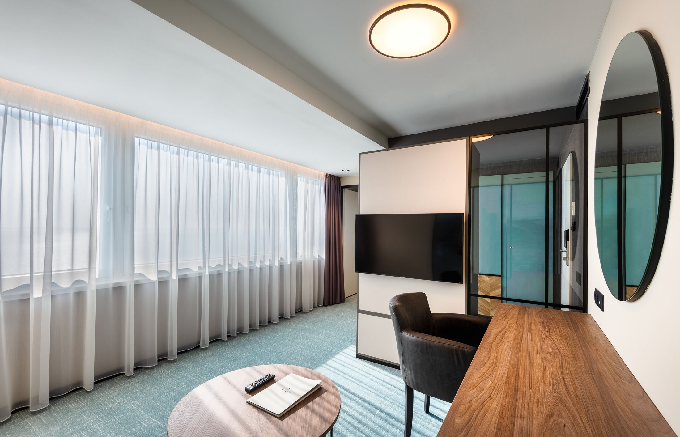 Een moderne hotelkamer met een groot raam, een flatscreen televisie, een houten bureau en een comfortabele stoel.