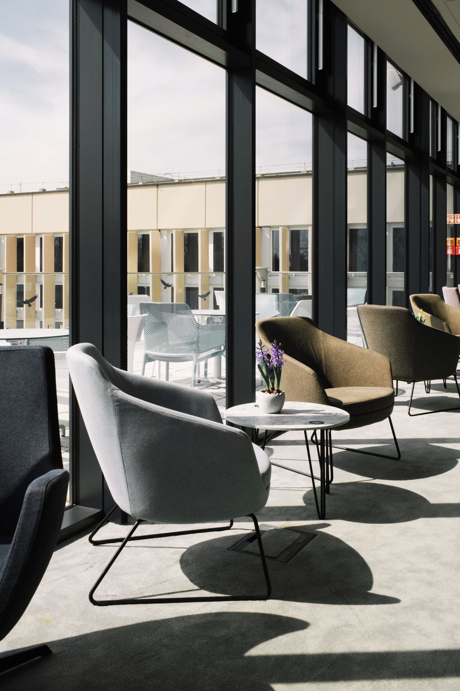Ein moderner Außenbereich mit modernen Stühlen, einem kleinen Tisch, großen Fenstern und Blick auf ein Gebäude im Hintergrund.
