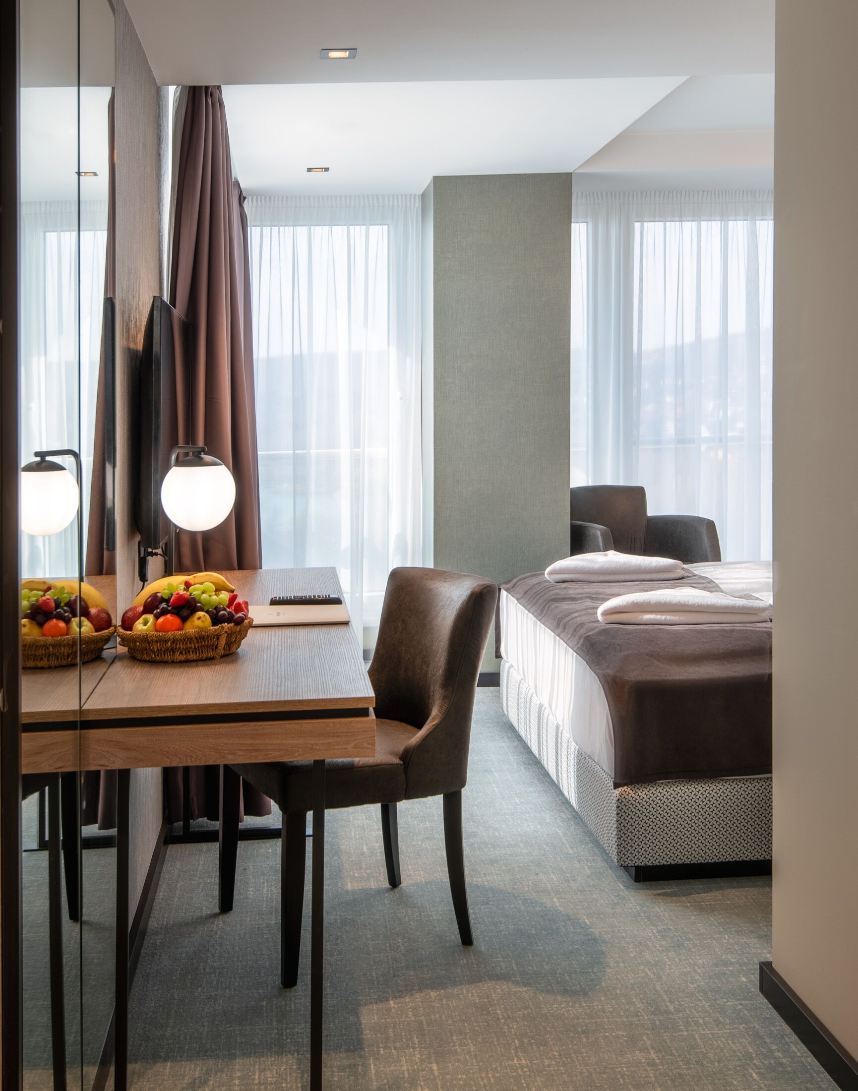 A hotel room with a bed, a desk with a chair, a fruit basket, and a large window with curtains.