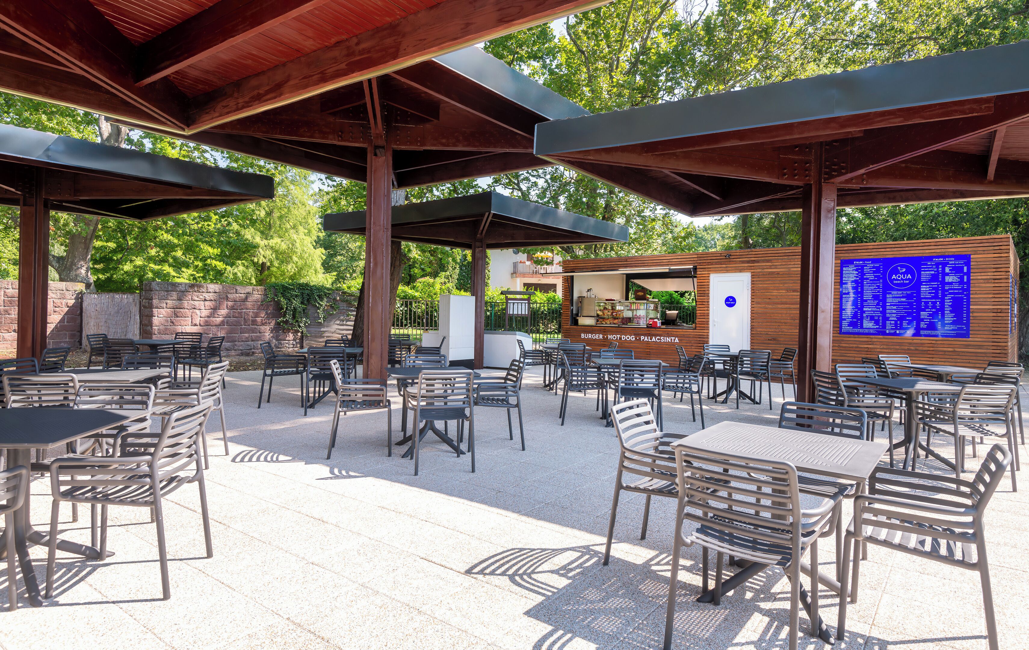 An outdoor patio with metal tables and chairs, a wooden structure overhead, and a menu board in the background.