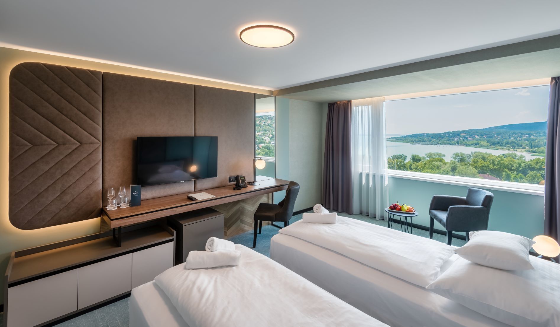 A hotel room with two beds, a desk with a TV, a view of the outdoors, and a balcony with a table and chairs.