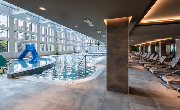 A modern indoor swimming pool with a blue slide, lounge chairs, and large windows.