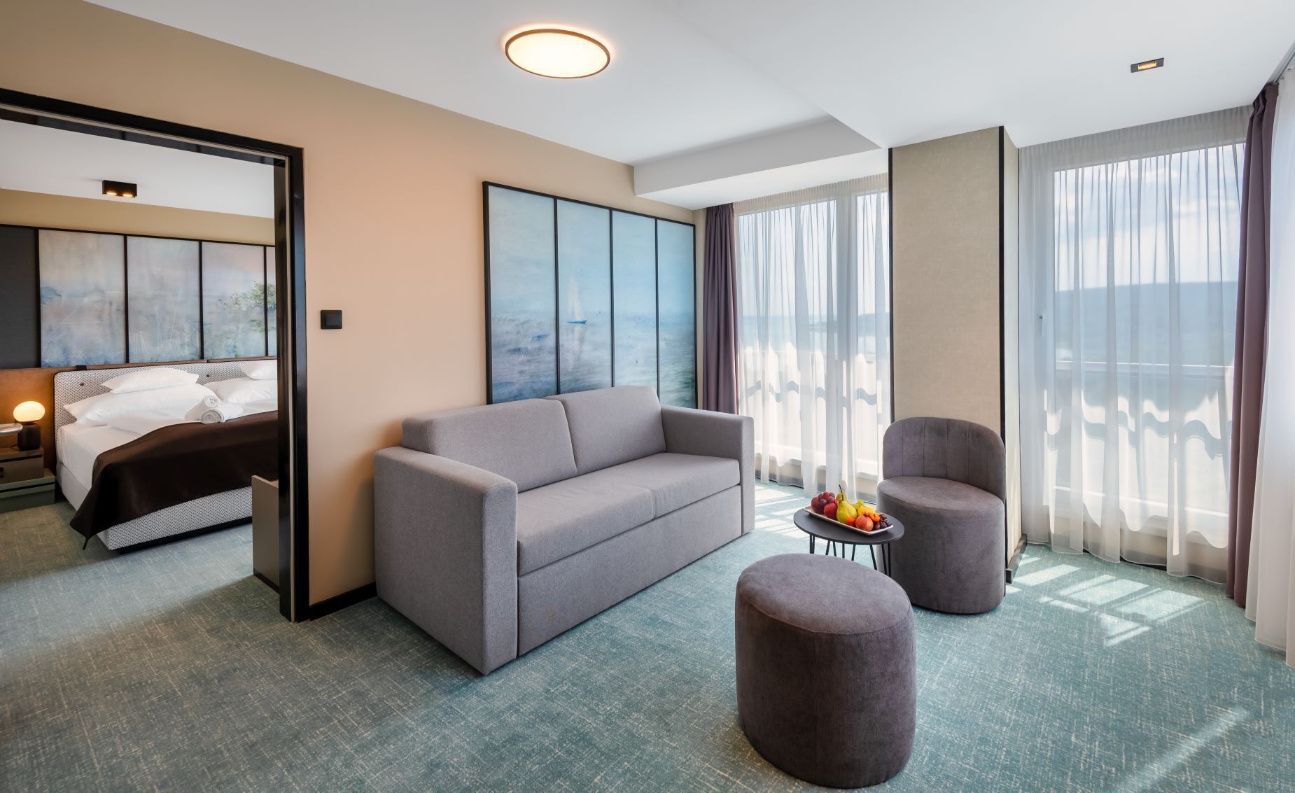 Una habitación de hotel moderna con una cama grande, un sofá gris, un otomanillo redondo y una mesita de noche con una lámpara.