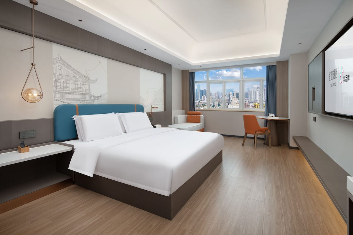 A modern hotel room with a large bed, a desk, and a chair. There is a window with a city view.