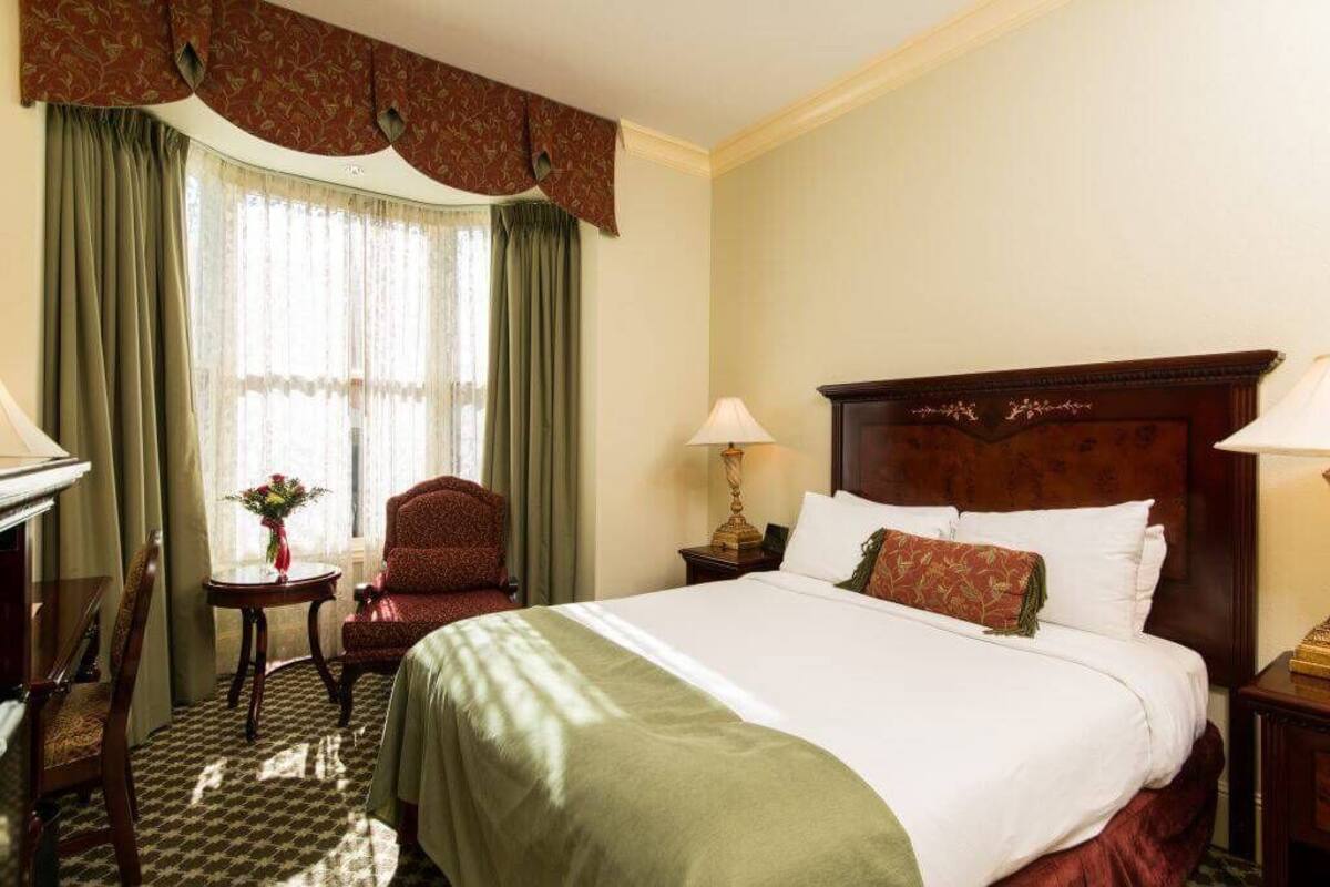 A hotel room with a large bed, a chair, a small table with a vase, and curtains.