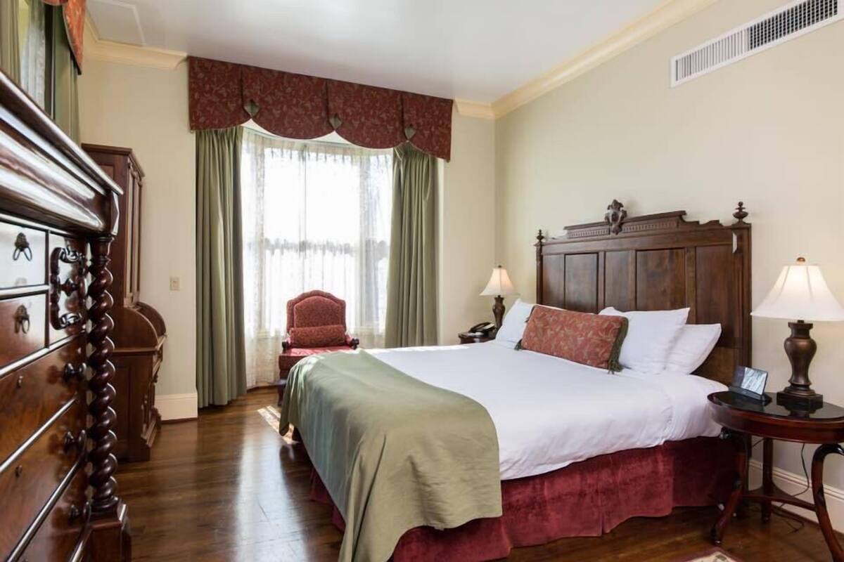 A hotel room with a large bed, wooden headboard, two bedside tables with lamps, a red armchair, and a wooden dresser.