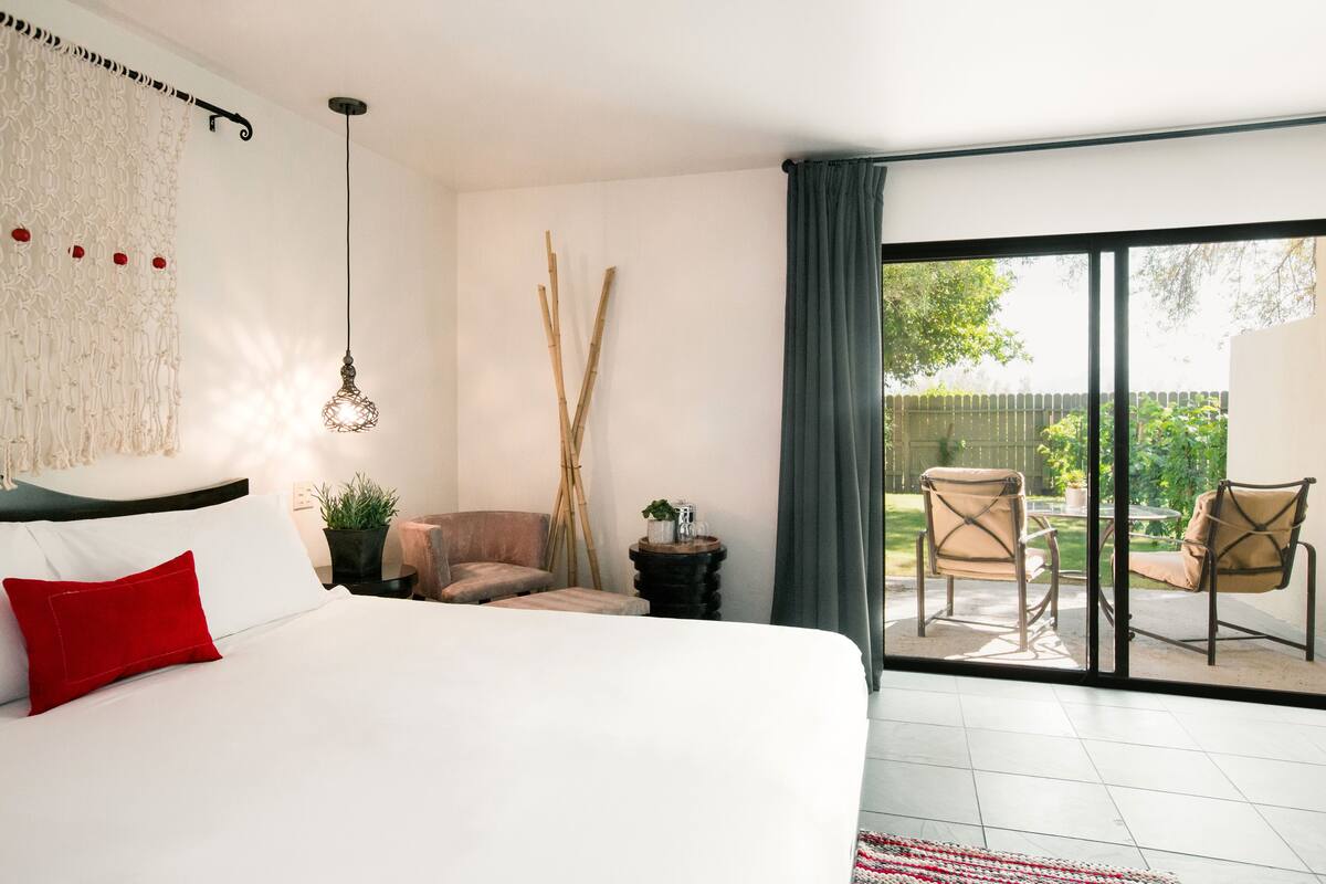 Une chambre à coucher avec un lit, un oreiller rouge, un luminaire suspendu, une petite table avec une plante, et une vue sur une terrasse extérieure avec des chaises et une table.