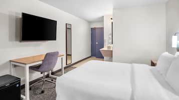 A hotel room with a bed, a desk with a chair, a television, a mirror, a bathroom with a sink and a towel dispenser.