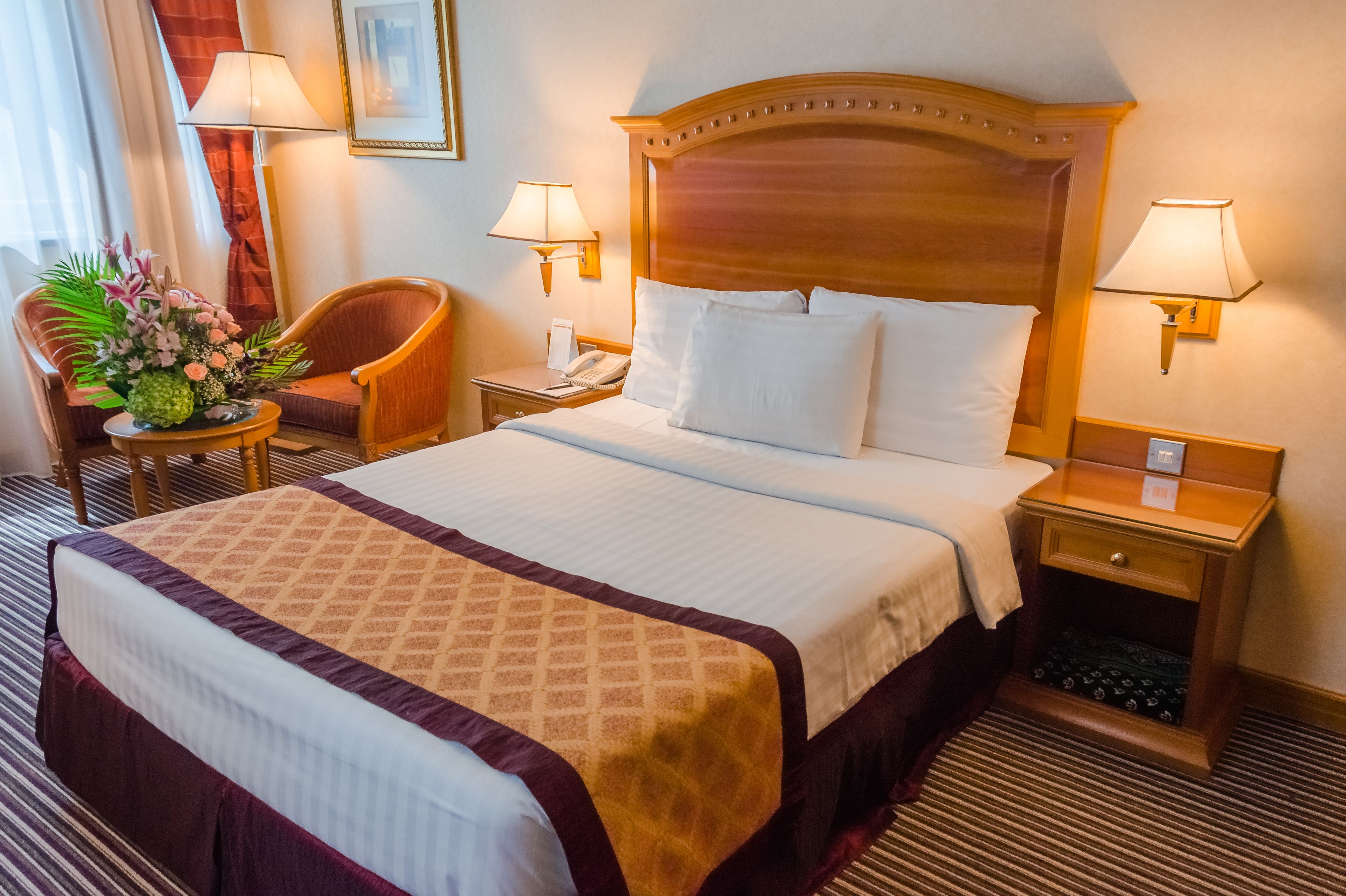 A hotel room with a large bed, two bedside tables, a chair, a small table with flowers, and two wall-mounted lamps.