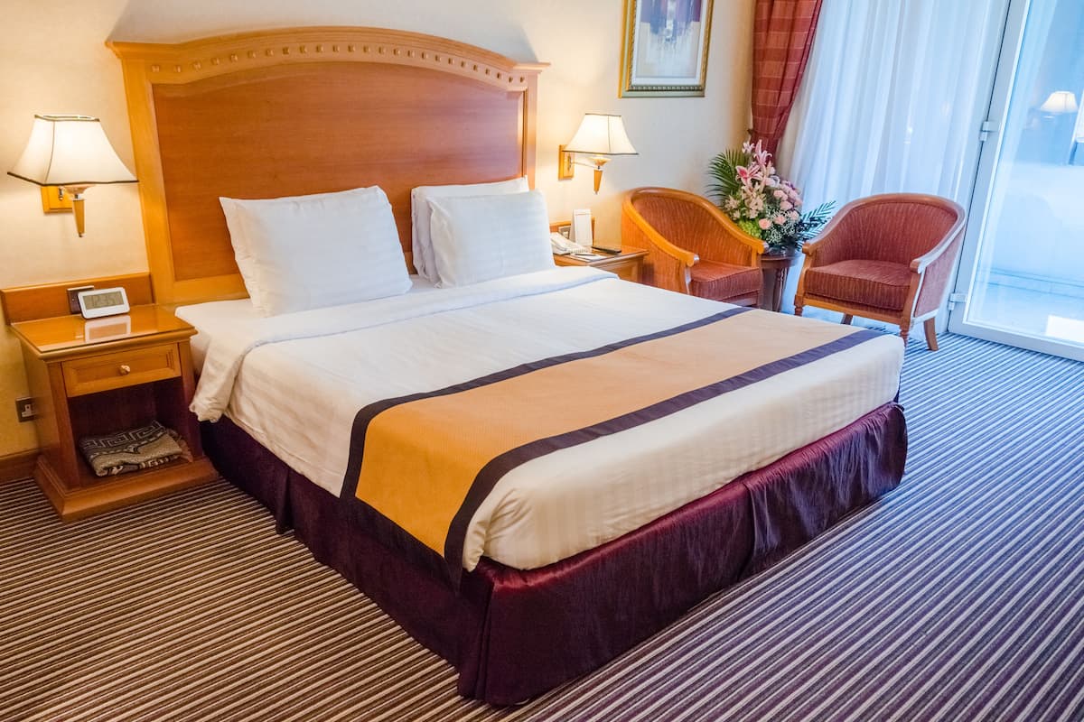 A hotel room with a large bed, two bedside tables, a nightstand, two lamps, a chair, and a potted plant.