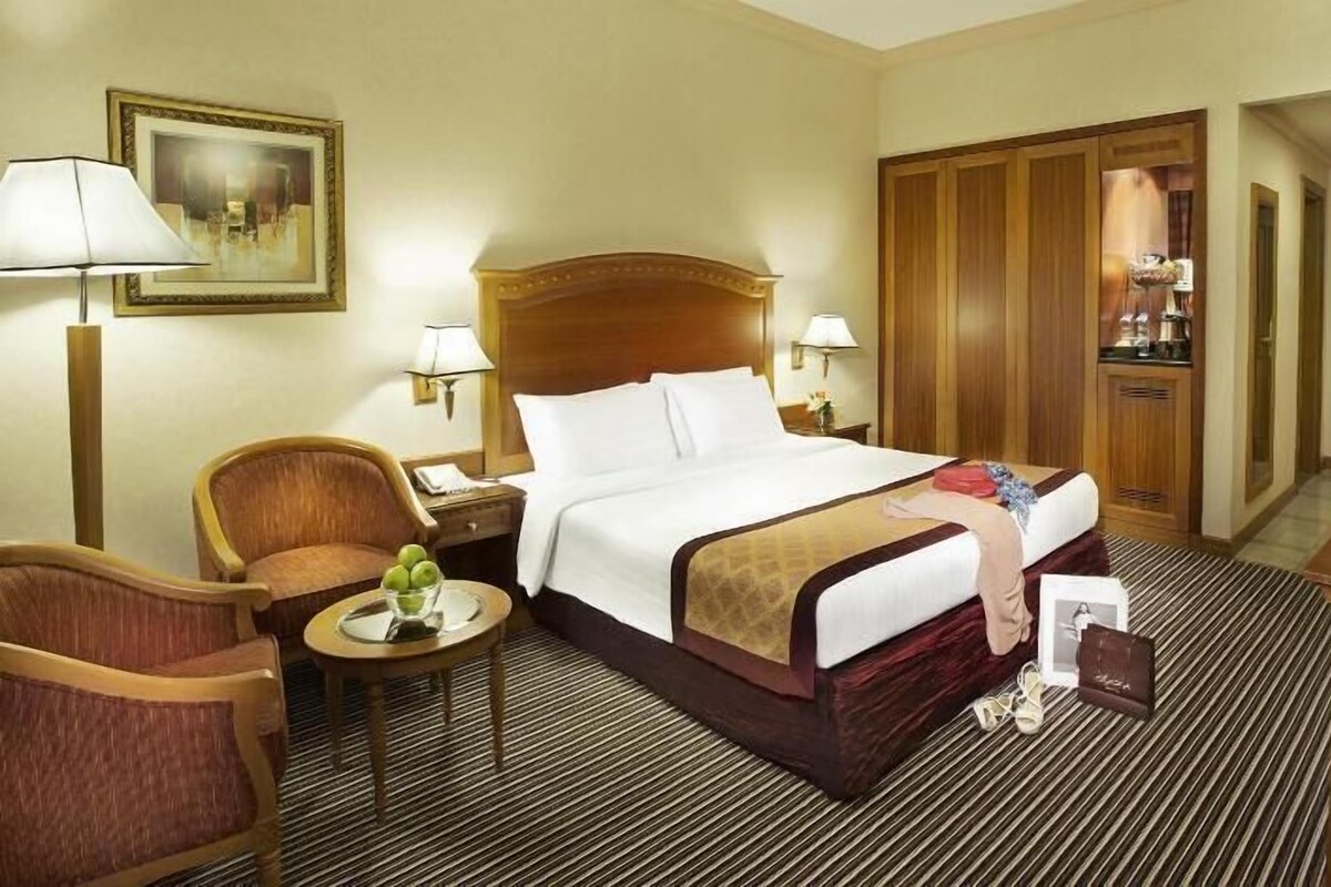 A hotel room with a bed, a chair, a small table with a vase of flowers, a lamp, a painting on the wall, and a television.