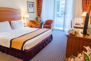 A hotel room with a large bed, two chairs, a desk with a computer, a lamp, and a window with curtains. - Avenue Hotel (Dubai)