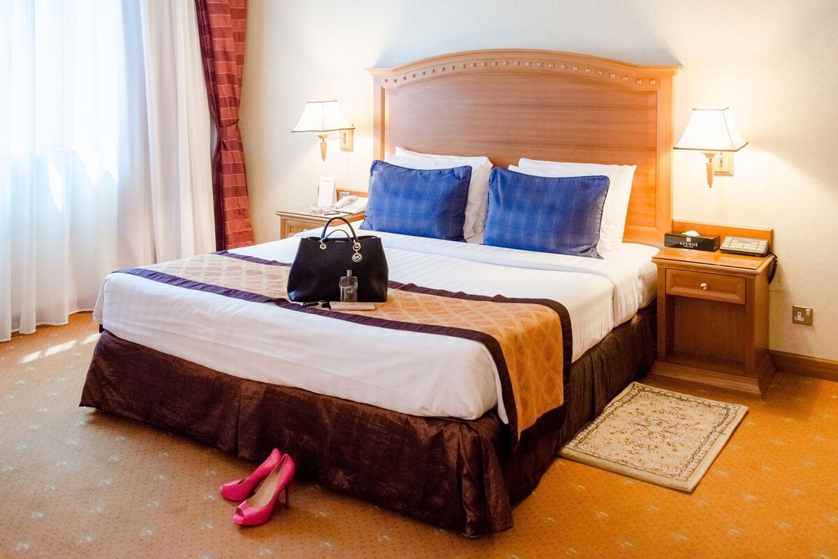 A hotel room with a large bed, two bedside tables, a nightstand, a lamp, a handbag, and a pair of shoes.