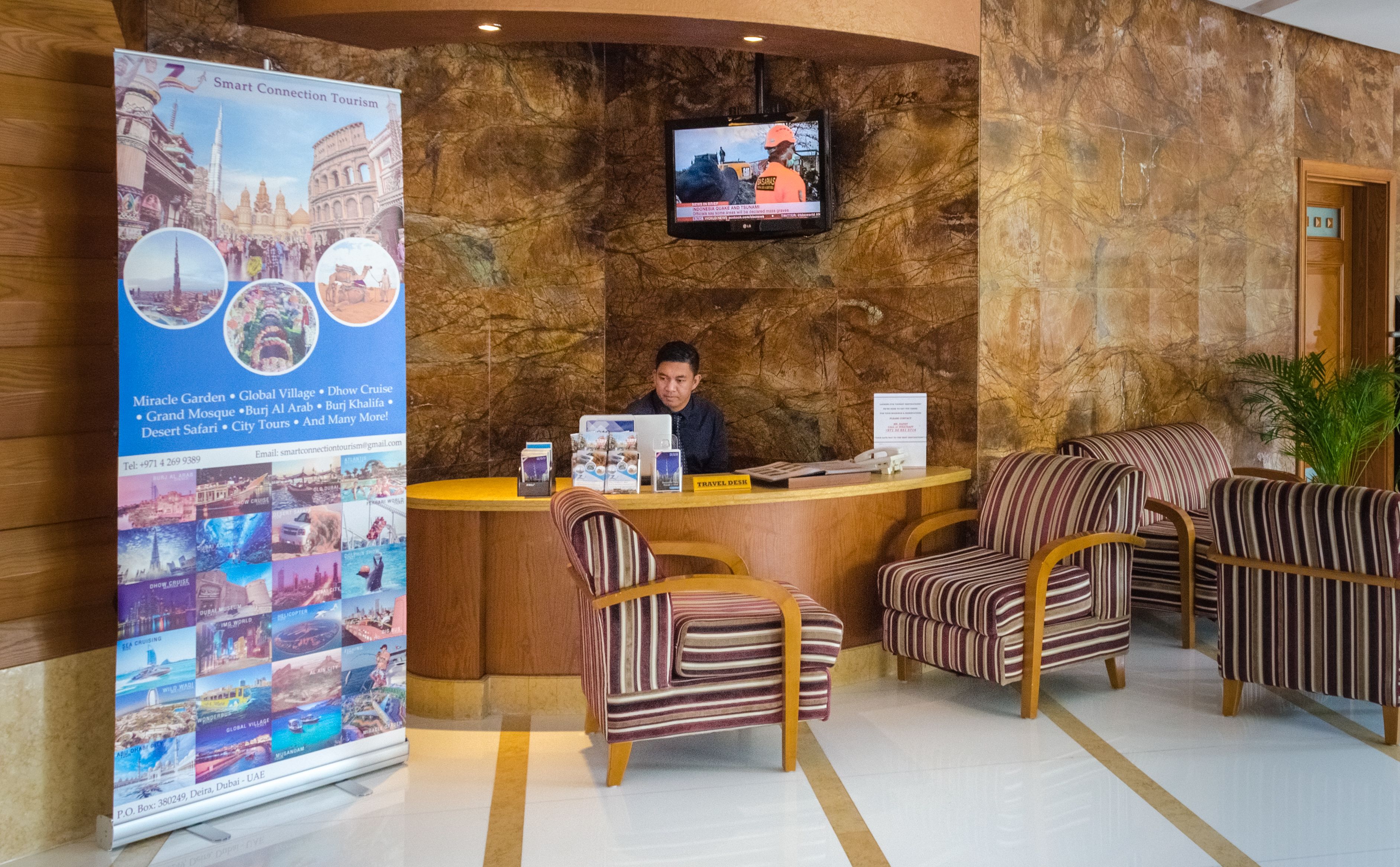 A reception area with a receptionist desk, two striped armchairs, a large poster with travel destinations, and a television mounted on the wall.