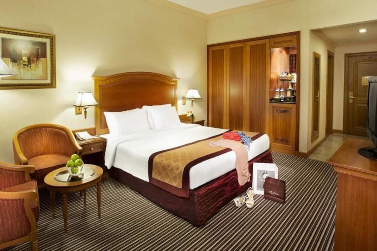 A hotel room with a bed, a chair, a small table with a fruit bowl, a TV, and a minibar.