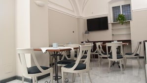 A dining area with white chairs and tables, a flat-screen TV mounted on the wall, and a potted plant on a shelf. - Hotel Centrum (Catania)
