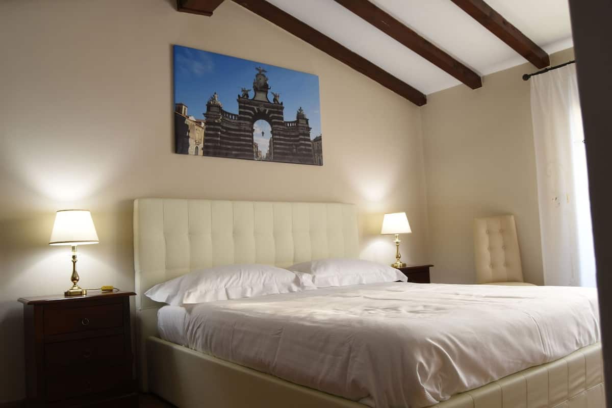 Habitación de hotel con una cama, dos lámparas de noche, una mesita de noche, una silla y un cuadro en la pared.