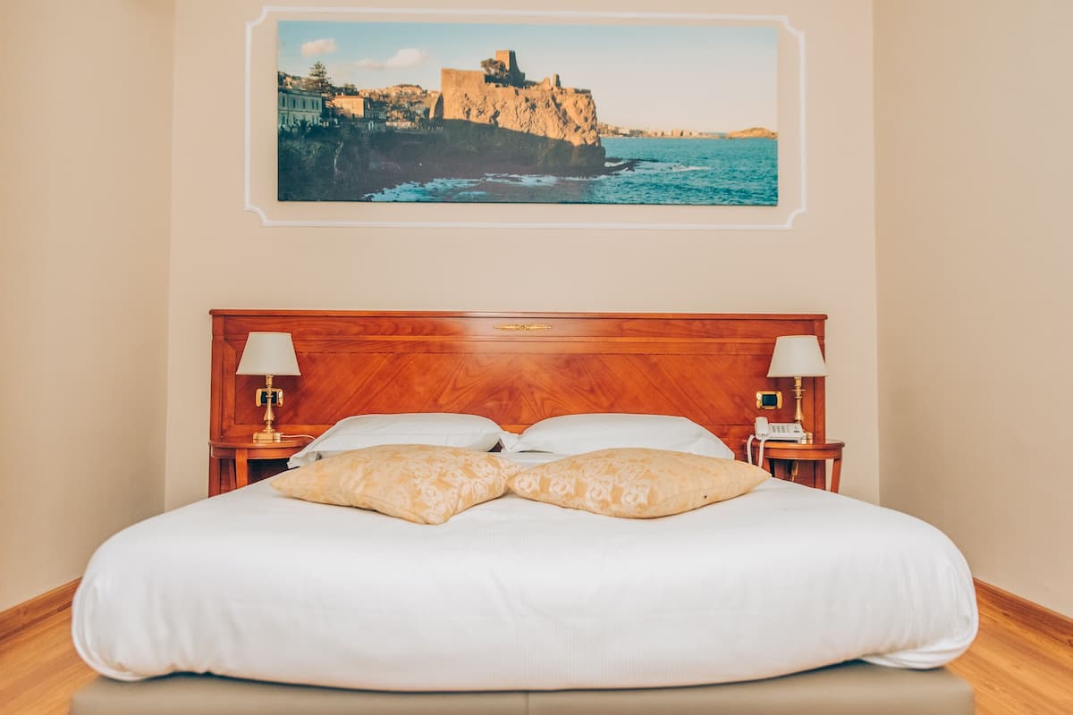 Una cama bien hecha con un cabecero de madera, dos lámparas de noche y un cuadro enmarcado de un paisaje costero.