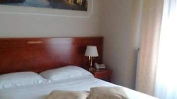 Quarto de hotel com uma cama grande, uma mesa de cabeceira com abajur e um quadro com vista urbana na parede.