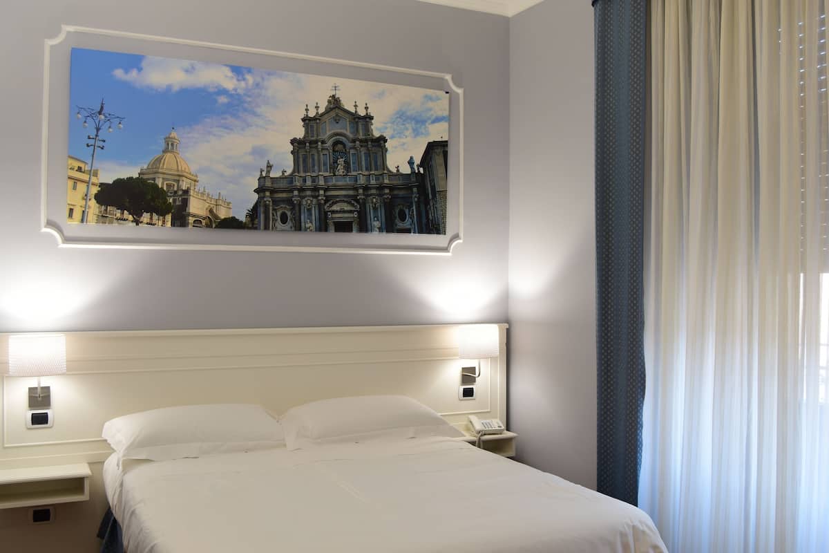 Una habitación de hotel con una cama, una mesita de noche, un teléfono y un cuadro de un edificio en la pared.