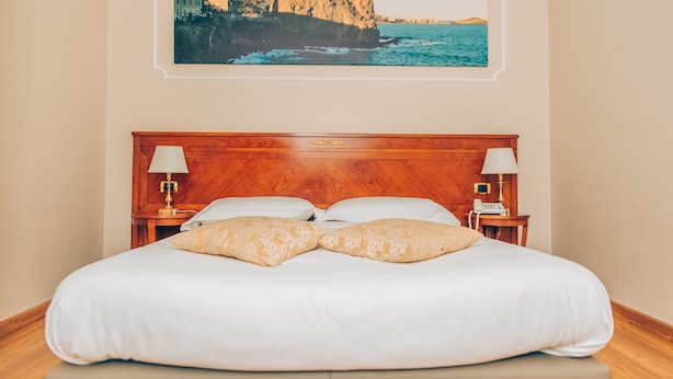 A neatly made bed with a wooden headboard, two bedside lamps, and a framed picture of a coastal landscape on the wall. - Hotel Centrum (Catania)