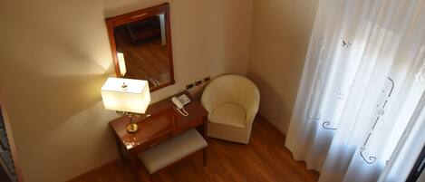 A small, well-lit room with a wooden desk, a chair, a bedside table with a lamp, a telephone, and a mirror.