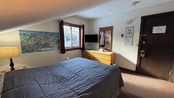 A bedroom with a bed, a dresser, a mirror, a window with curtains, a wall-mounted TV, and a door with a sign.
