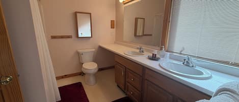 Une salle de bain comprenant des toilettes, deux lavabos, un miroir et une fenêtre avec des stores.